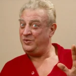 Rodney Dangerfield adjusting his red tie, delivering his famous "no respect" routine on stage.