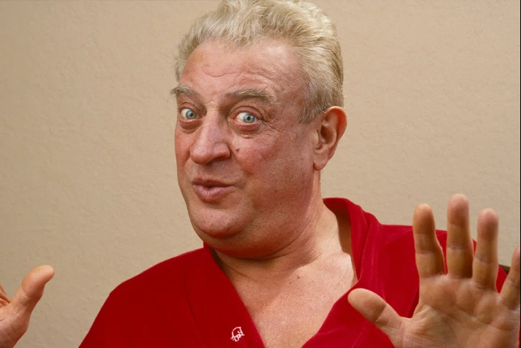 Rodney Dangerfield adjusting his red tie, delivering his famous "no respect" routine on stage.